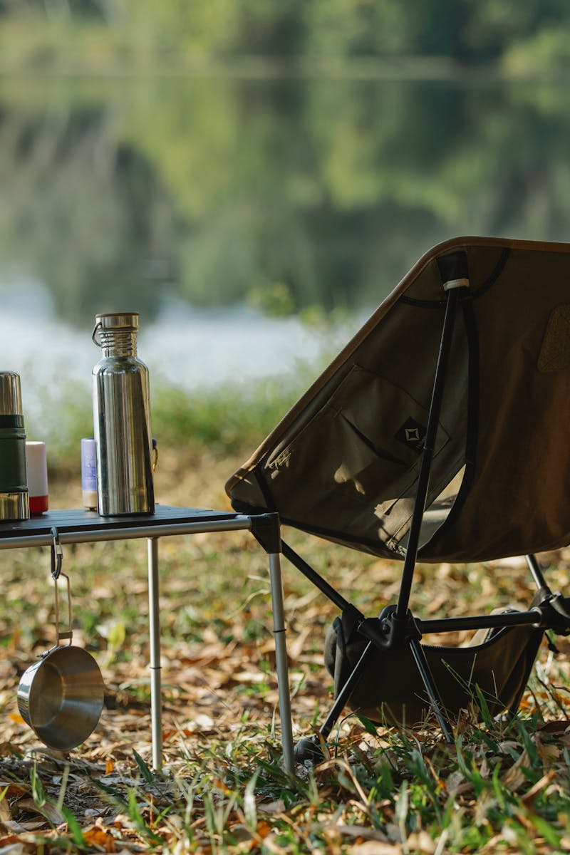 A peaceful lakeside campsite with a folding chair, table, thermoses, and saucepan.